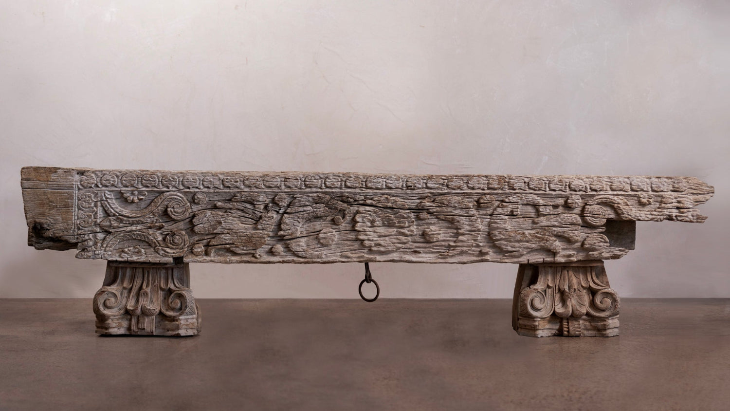 An antique carved wooden table or bench with ornate scrollwork details, featuring a rustic weathered finish and a hanging ring on the front, set against a neutral studio backdrop.